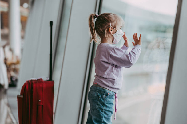 Toddler girl at airport in face mask. Child look at airplane after coronavirus outbreak. Safe travel and flying with child in virus pandemic. Kid at departure gate. Vacation after covid-19 lockdown. - Foto, afbeelding