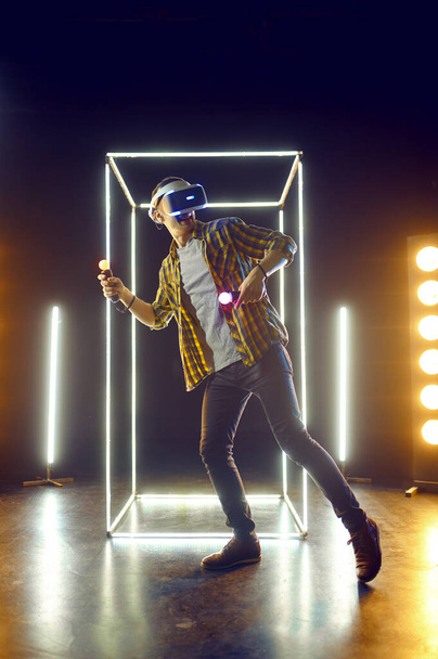 Man gaming in virtual reality headset and gamepad in luminous cube. Dark playing club interior, spotlight on background, VR technology with 3D vision - 写真・画像