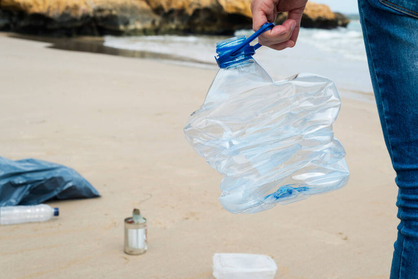 gros plan d'un jeune homme caucasien ramassant une bouteille en plastique usagée dans le sable d'une plage isolée, comme une action pour nettoyer l'environnement naturel - Photo, image