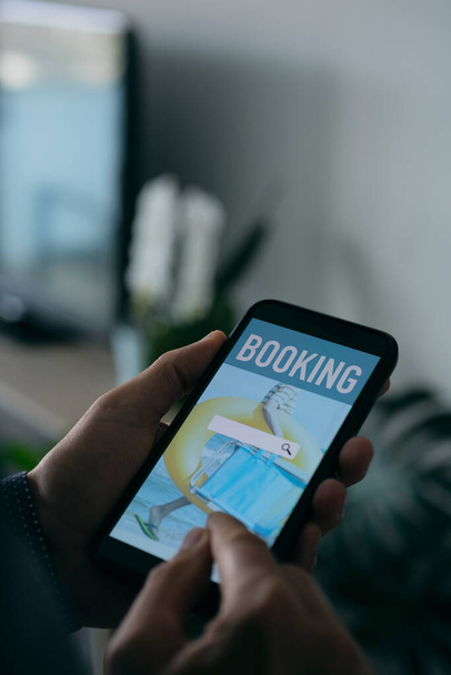 un jeune homme caucasien réserve ses vacances en ligne avec son smartphone, avec un moteur de recherche simulé dans son écran, debout à son salon - Photo, image