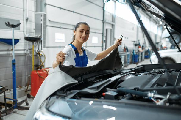 Female mechanic stands at the hood, car service. Vehicle repairing garage, woman in uniform, automobile station interior on background - 写真・画像