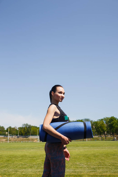 Young slim girl in sport clothes with yoga mat on stadium green lawn before training. Sport, healthy way of life, fitness concept. - Foto, afbeelding