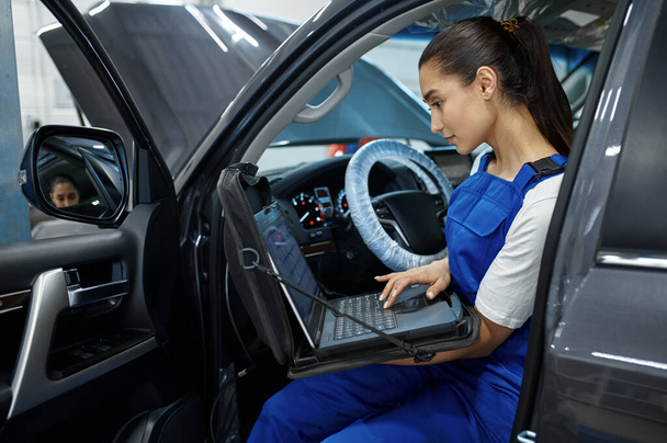 Female mechanic with laptop does the engine diagnostics, car service, professional inspection. Vehicle repairing garage, woman in uniform, automobile station interior on background - Photo, Image