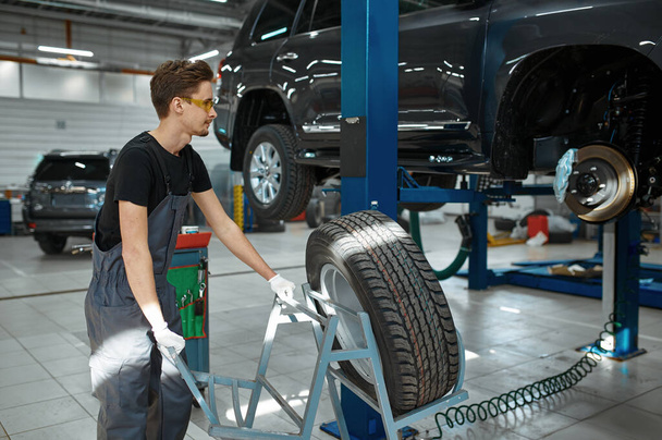 Male mechanic fixes problem with wheel, car service. Vehicle repairing garage, man in uniform, automobile station interior on background - Photo, Image