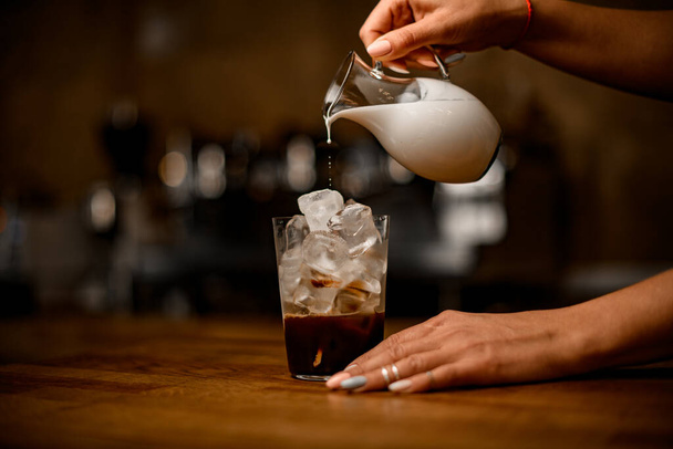 hand holds jug of milk and pours it into glass with coffee and ice - Фото, зображення