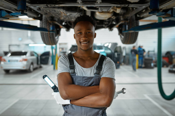 Male worker checks car suspension, auto service. Vehicle repairing garage, man in uniform, automobile station interior on background - Foto, imagen