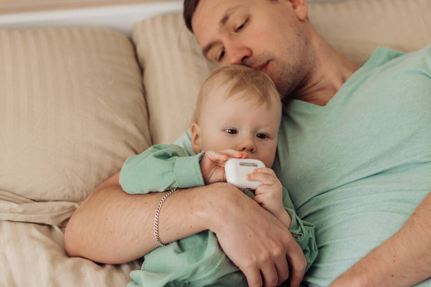 Young happy father is lying on the bed with his baby son in his arms - 写真・画像
