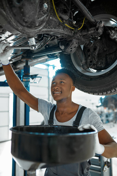 Male mechanic drains the oil, car service. Vehicle repairing garage, man in uniform, automobile station interior on background - Foto, Imagen