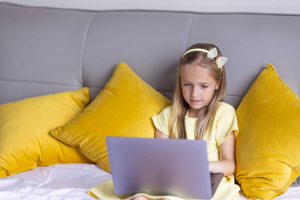 Cute little caucasian girl with blonde hair in fashionable dress illuminating yellow color sitting at home during coronavirus pandemic quarantine and using laptop. Stay at home during covid-19 - Фото, зображення