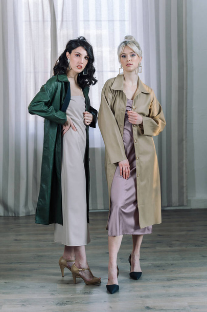 Young girls stands in the studio on the background of curtains. Fashionable modern raincoats are worn over the dresses. Beautiful healthy teeth are visible. - Foto, afbeelding