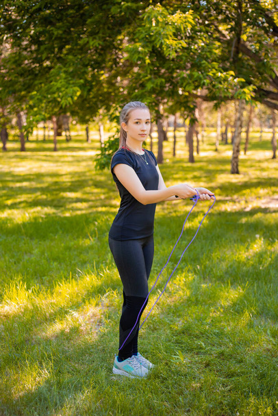 夏の公園の芝生の上には、スキップロープを手にした若い女性が立っている。健康的なライフスタイルと屋外運動の概念. - 写真・画像
