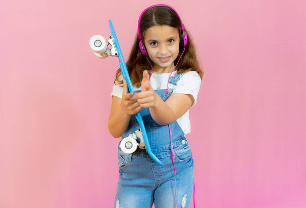 Lächelndes kleines Mädchen mit Kopfhörern und Skateboard auf rosa Hintergrund - Foto, Bild