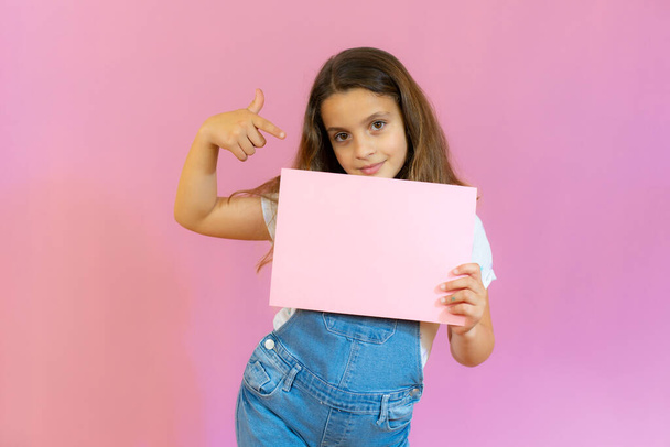 Smiling little girl holding empty pink board isolated over pink background. - 写真・画像