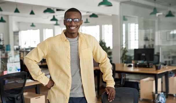 Portrait of a young African businessman smiling while leaning on a chair by his desk in an office after hours - Photo, Image