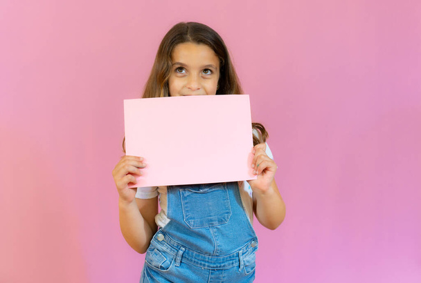 усміхнена дівчинка тримає порожню рожеву дошку ізольовано на рожевому фоні
. - Фото, зображення