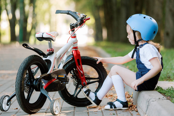 Cheerful smiling girl in helmet sitting near bicycle. Kid checking bike tires  - Foto, immagini
