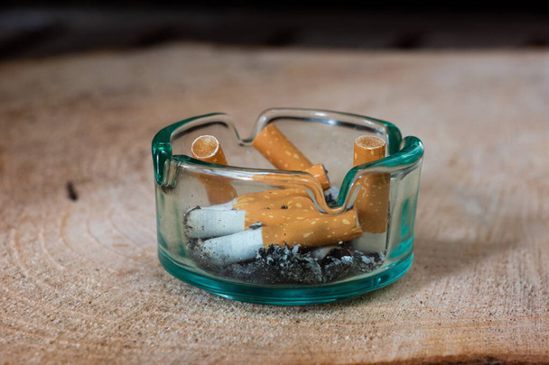 Cenicero de cigarrillo de vidrio transparente con colillas y ceniza en tablero de madera. - Foto, Imagen