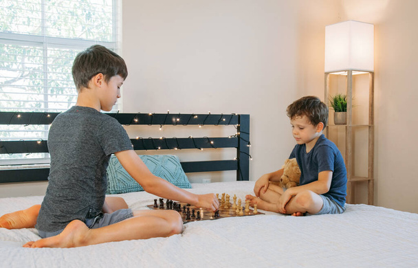 Два хлопці грають в шахи вдома на ліжку. Друзі розважаються. Діти практикують гру на дошці
 - Фото, зображення