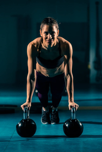 A young woman in a gym doing push-ups with the help of a ball-shaped weight known as a kettlebell. - Photo, Image