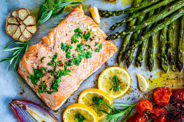 Sheet pan dinner - roasted salmon steak with asparagus, lemon ,rosemary, tomatoes, onion and garlic on cooking pan on wooden table  - Photo, image