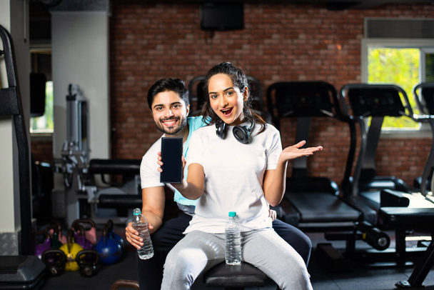 Sportif indien asiatique homme et femme reposant entre les exercices et en utilisant smartphone à la salle de gym, présenter quelque chose ou prendre selfie photo - Photo, image