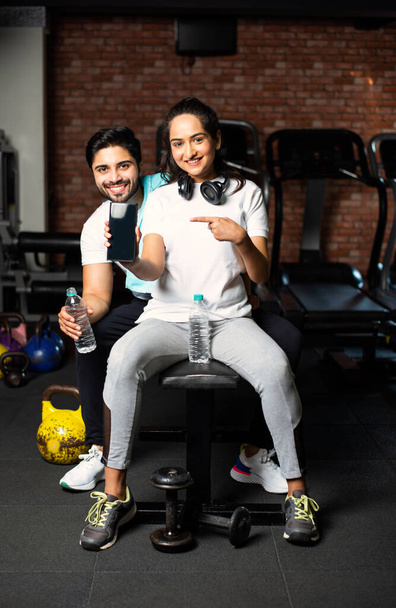 Sporty Indian asian man and woman resting between exercises and using smartphone at gym, presenting something or taking selfie picture - Photo, Image