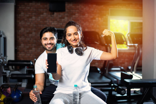 Sportovní indické asijské muž a žena odpočívající mezi cvičeními a pomocí smartphone v tělocvičně, prezentaci něco nebo focení selfie - Fotografie, Obrázek