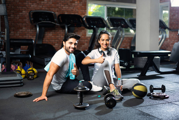 Indiaas jong paar in de sportschool rusten of pauze nemen van lichaamsbeweging, drinkwater, het tonen van positieve uitdrukkingen - Foto, afbeelding