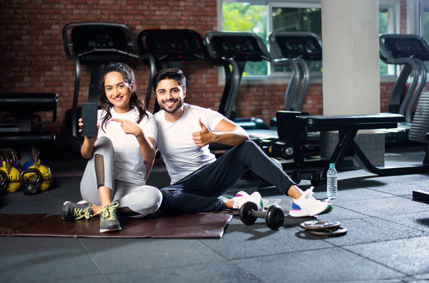 Sporty indiano asiatico uomo e donna riposo tra esercizi e utilizzando smartphone in palestra, presentando qualcosa o scattando foto selfie - Foto, immagini