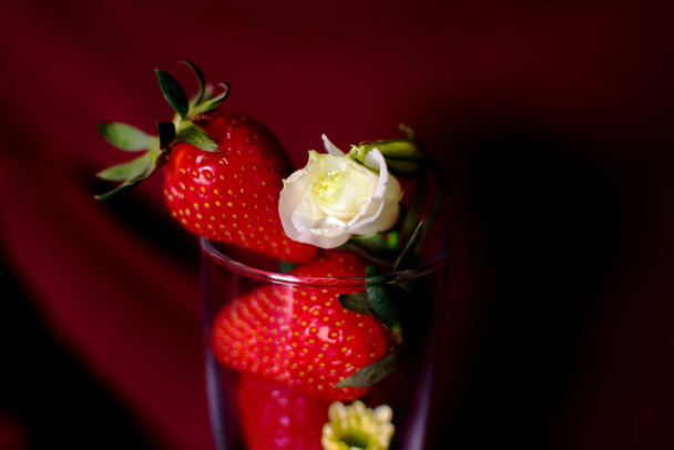 バラのあるガラスの中のイチゴ。バラの上に水の滴です。写真はブルゴーニュを背景に撮影。選択的焦点で撮影した写真. - 写真・画像