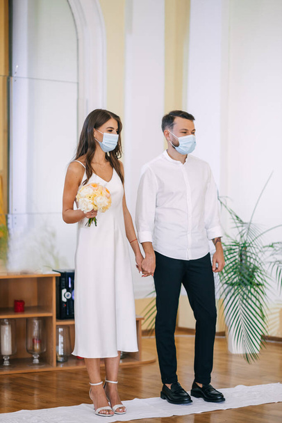 a couple of newlyweds in the registry office. obtaining an official marriage certificate. The groom in a shirt and trousers and the bride in an elegant white dress wearing medical masks. A wedding during a pandemic. - Foto, imagen