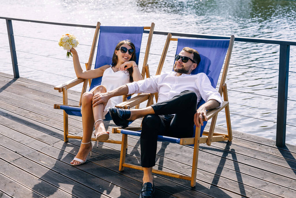 recém-casados jazem em espreguiçadeiras junto à piscina. Uma mulher em um vestido de luz branca com alças de espaguete e um homem em uma camisa e calças em óculos de sol. - Foto, Imagem