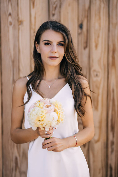 Een mooie meisje-bruid in een witte trouwjurk, met een delicaat boeket pioenrozen staat tegen de achtergrond van een houten muur. Stijlvolle kapsel en mooie make-up. - Foto, afbeelding