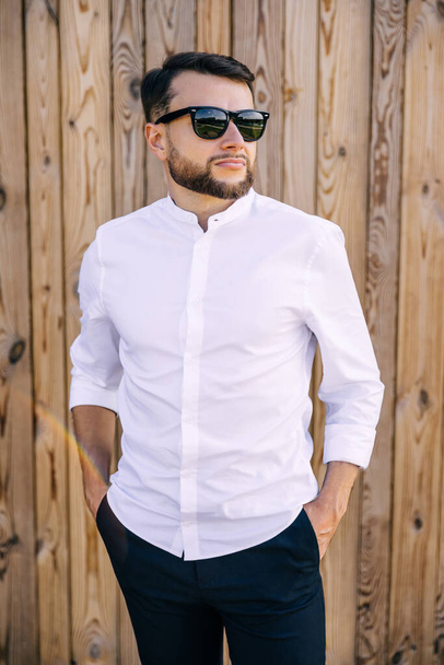 ein junger gutaussehender, modischer Mann mit Brille steht vor einer Holzwand, hält seine Hände in der Tasche. - Foto, Bild