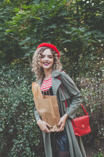 Šťastná hezká žena s kudrnaté vlasy v červeném baretu držení řemeslné papírové tašky s francouzskou bagetou venku - Fotografie, Obrázek