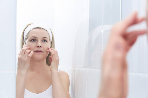 bela mulher de meia-idade faz auto-massagem do rosto em torno dos olhos na frente de um espelho  - Foto, Imagem