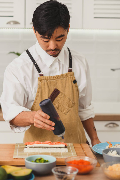 Портрет азиатского шеф-повара, делающего суши. Процесс суши-ролла добавляет соус к суши - Фото, изображение