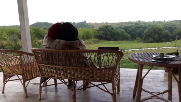 Zamilovaný pár sedí na terase na třtinové lavičce, objímají se a přikrývají dekou - Záběry, video