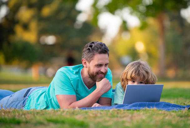 Ojciec uczy syna korzystać z laptopa, tata i chłopiec szkolny wygląd ekranu komputera i tabletu. - Zdjęcie, obraz