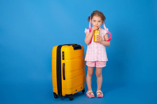 Забавная девочка в розовой одежде держит стакан апельсинового сока с желтым чемоданом на синем фоне. - Фото, изображение