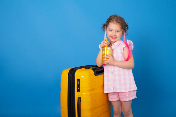 Funny child girl in pink clothes holding glass of orange juice with yellow suitcase on blue background. - Foto, Imagen