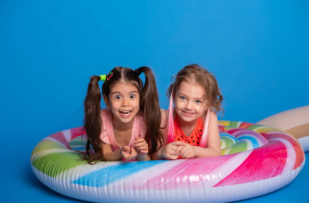 two happy child girl in swimming suit lying on colorful inflatable mattress lollipop on blue background - Фото, изображение