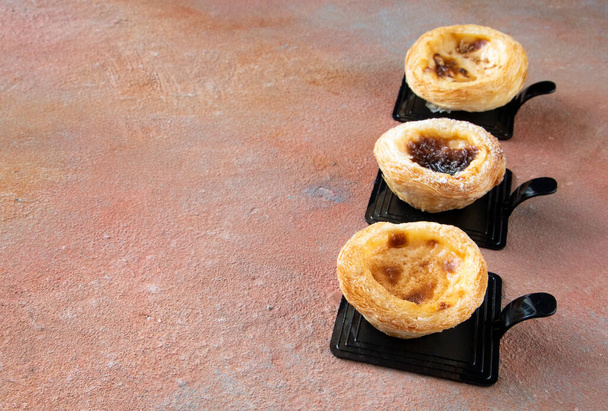 Traditional dessert from Portugal - Pastel de nata made of puff pastry, egg yolks, and cinnamon on black plate with copy space on a beautiful background reminiscent of the Martian surface - Photo, Image