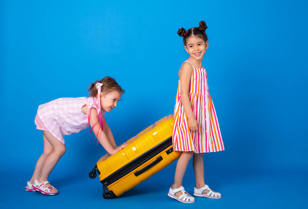 hauska pikku tytöt ystäviä värikkäissä vaatteissa kuljettaa keltainen matkalaukku sinisellä pohjalla. - Valokuva, kuva