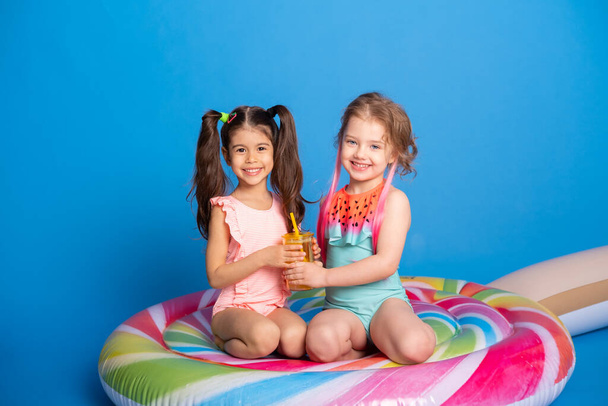 two happy child girl in swimming suit drinking orange juice sitting on colorful inflatable mattress on blue background - Photo, Image