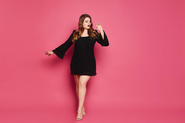 Plus size model girl in black gown makes step up on the pink background - Foto, immagini