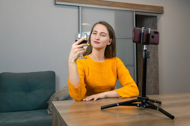 Νεαρή μελαχρινή blogger με πορτοκαλί μπλούζα και ένα ποτήρι κόκκινο κρασί κάθεται μπροστά από ένα τρίποδο με ένα κινητό τηλέφωνο σε ένα κομψό εσωτερικό χώρο. Γυναίκα blogger κρασιού - Φωτογραφία, εικόνα