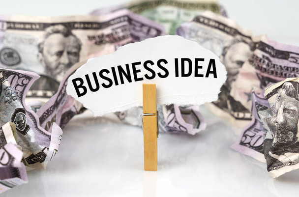 Επιχειρηματική και οικονομική έννοια. Υπάρχουν δολάρια στο τραπέζι και υπάρχει ένα μανταλάκι με χαρτί στο οποίο είναι γραμμένο - BUSINESS IDEA - Φωτογραφία, εικόνα