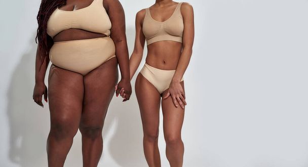 Cropped shot of two african american women in underwear with different body size holding each other hands, standing together isolated over gray background - Foto, afbeelding