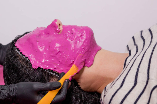 Alginate mask application process. A female hands of a beautician holding spatula. The procedure for skin care in the beauty salon. Woman lying on a couch in the office of a beautician - Photo, Image
